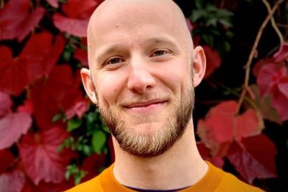 A head and shoulders shot of a smiling man with a shaved head and beard, wearing an orange jumper.