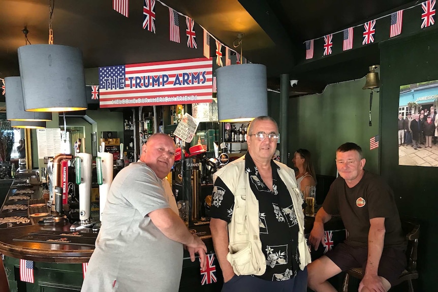 Three men lean on a a bar inside a pub with a banner saying 'The Trump Arms - welcome our American friends' behind them