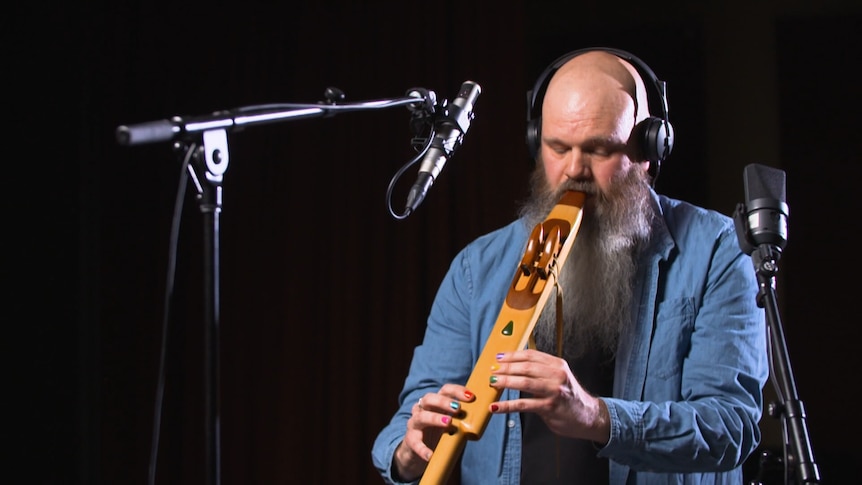 Adam Page plays harp flute with his eyes closed. He wears headphones and plays in front of two microphones.