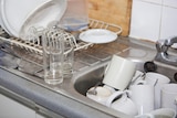 A  stainless steel sink full of dirty coffee cups and dishes, with a dish rack and glasses on the left-hand side.