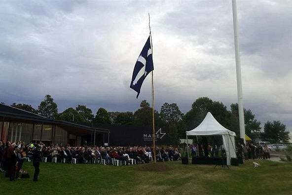 A group of people celebrate the Eureka Stockade's 160th anniversary in Ballarat, with the Eureka flag flying above them.