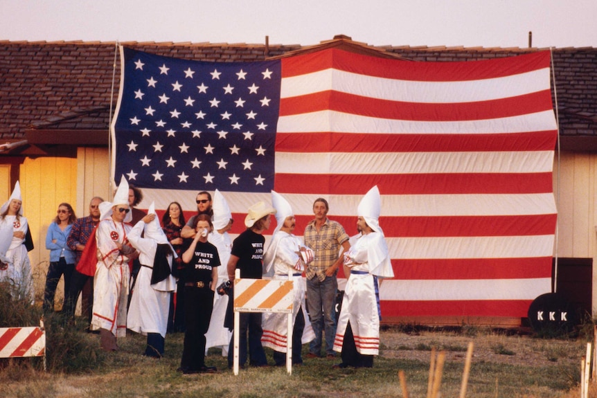 Robed clan members stand in front of an American flag attached to a house during a KKK meeting in Atlanta.