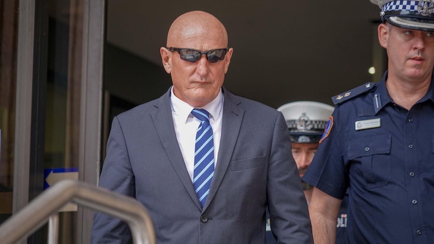 A bald man wearing a suit and sunglasses, leaving court flanked by police officers in uniform