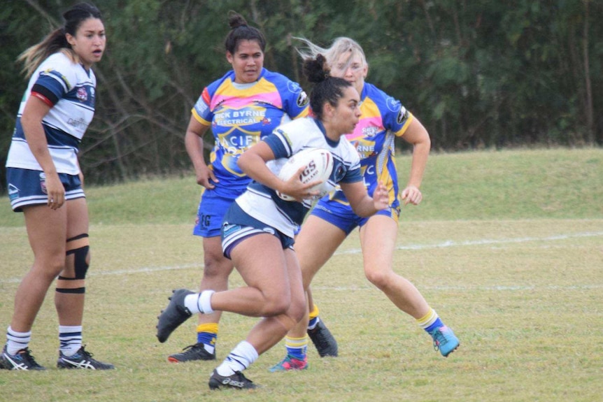 A woman in a blue and white footy uniform runs with the ball with three women to her side.