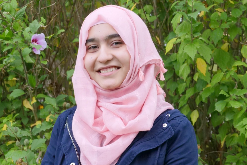 A teenage girl smiling wearing a pink hijab in front of a flowering tree.