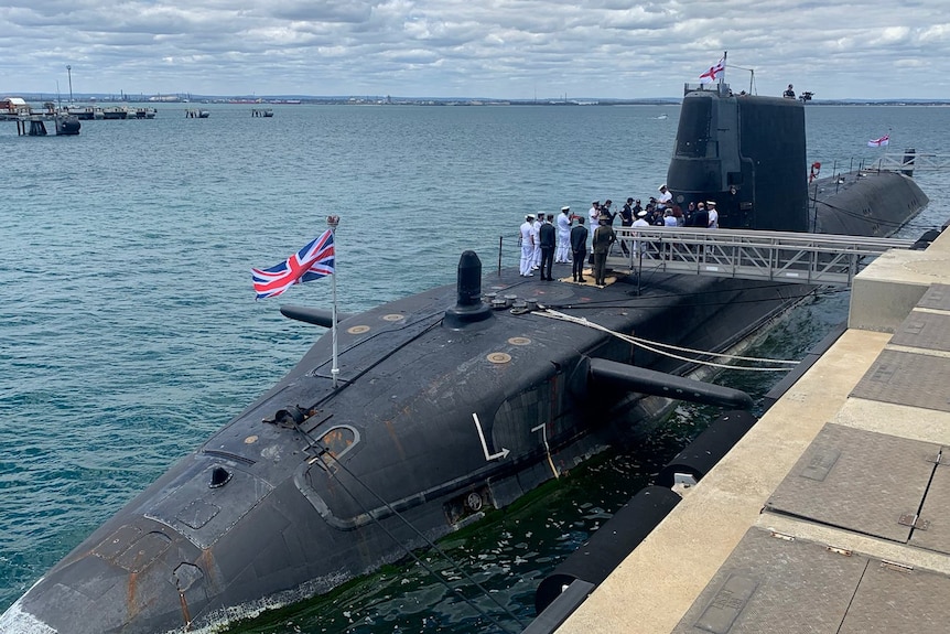 A submarine with a British flag and navy personnel on the top docked at a wharf with the ocean in the background.