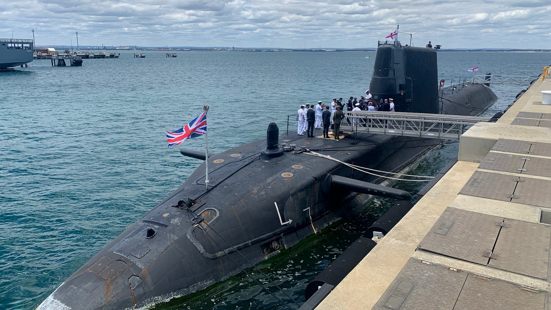 一艘悬挂英国国旗的潜艇和顶部的海军人员停靠在一个码头，背景是大海。