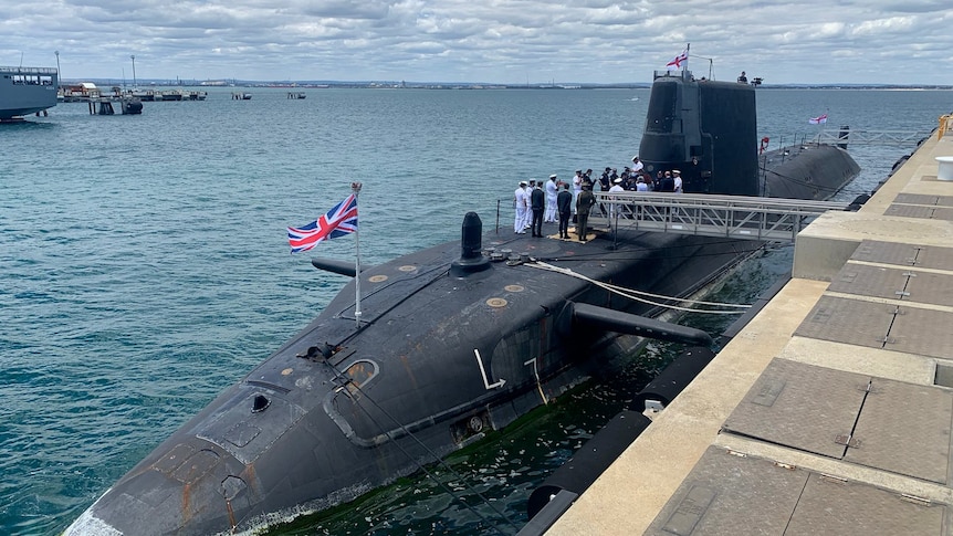 A submarine with a British flag and navy personnel on the top docked at a wharf with the ocean in the background.