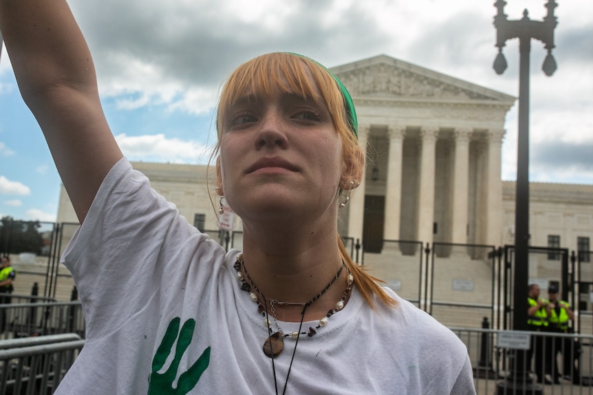 A woman with red hair and green bandana stands in front of the Supreme Court with her first in the air