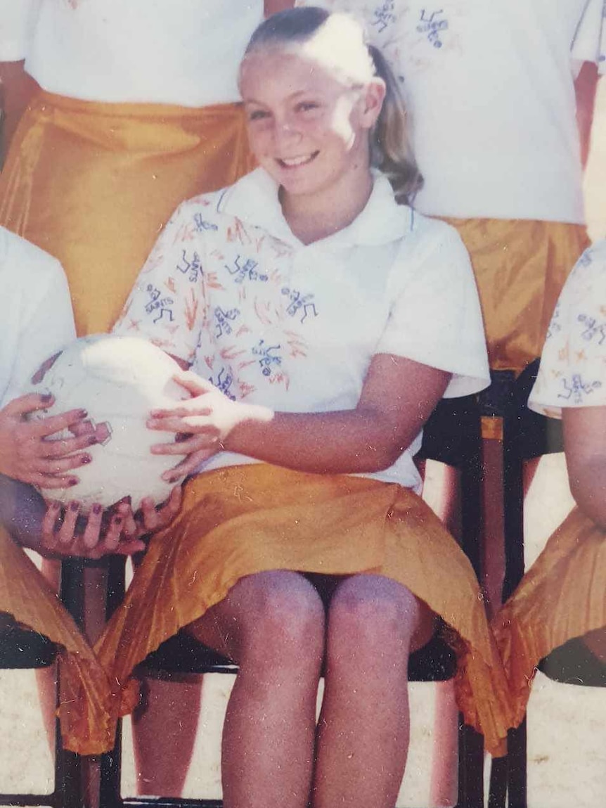 A young girl in white top and yellow skirt sitting down holding a netball 