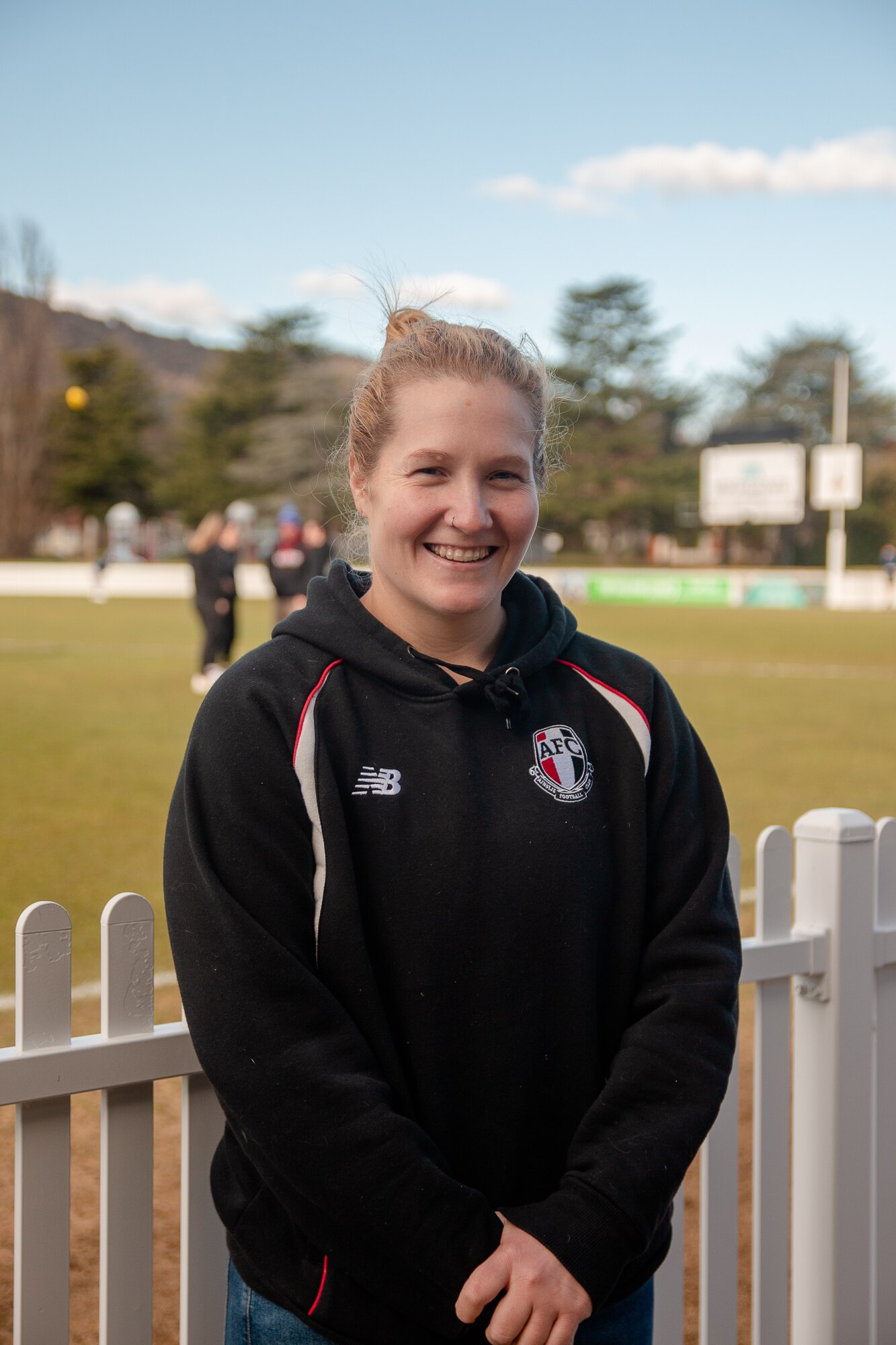Britt Tully stands in front of a white picket fence at a football oval and smiles.