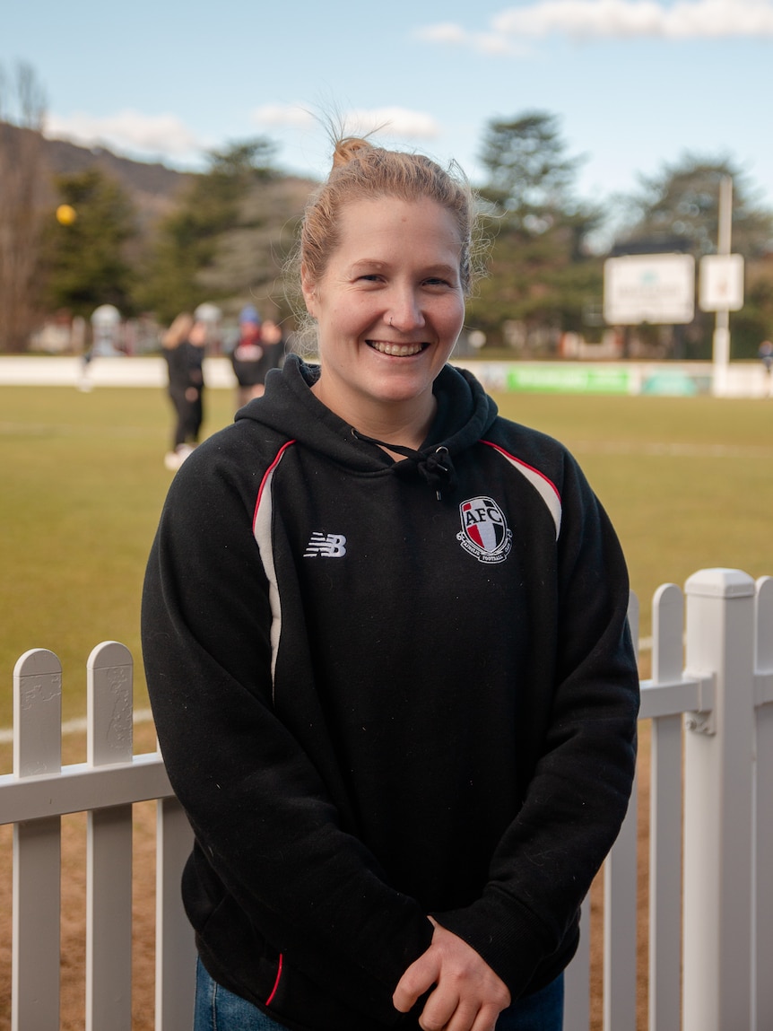 Britt Tully stands in front of a white picket fence at a football oval and smiles.