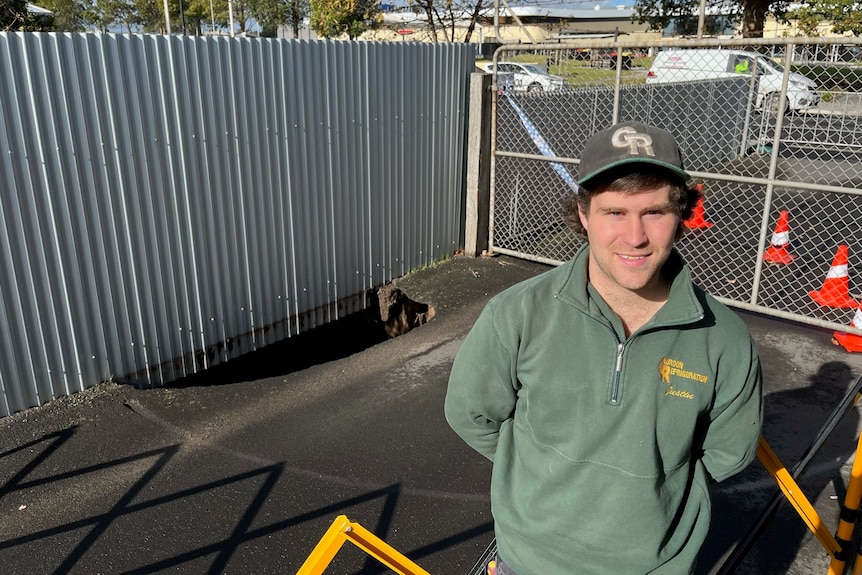 A man wearing a green jumper standing in a driveway next to a hole under a fence