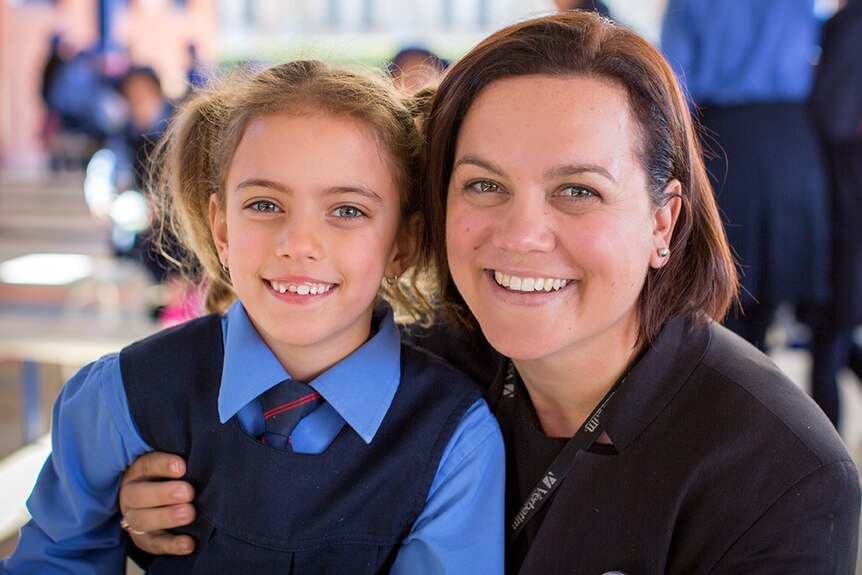 A smiling young girl in school uniform pinafore with tie, has face pressed close to a smiling woman, with shoulder-length brown.