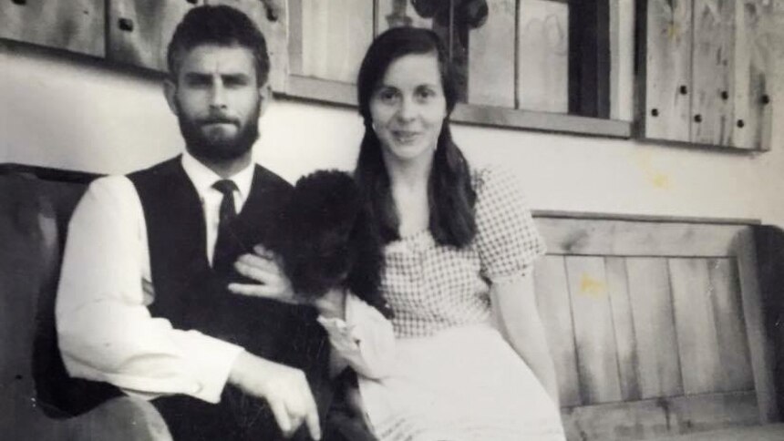 A black and white photo of a young man and woman sitting on a wooden bench, holding a black dog.