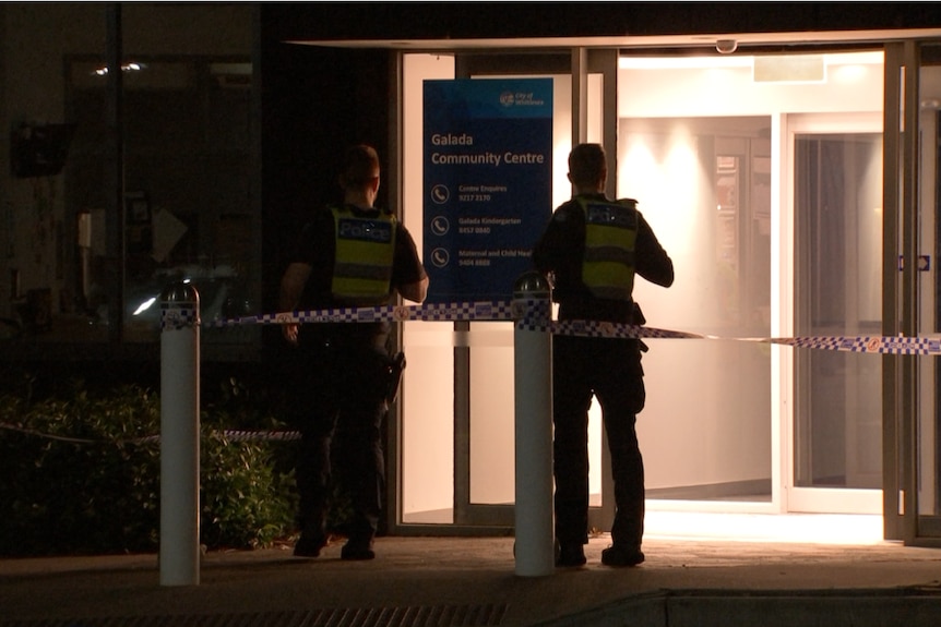 Two police are silhouetted as they stand at night outside a dimly lit community centre building.