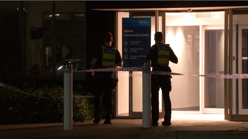 Two police are silhouetted as they stand at night outside a dimly lit community centre building.
