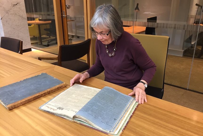 Photograph of an older white woman reading from a bound collection of old newspapers