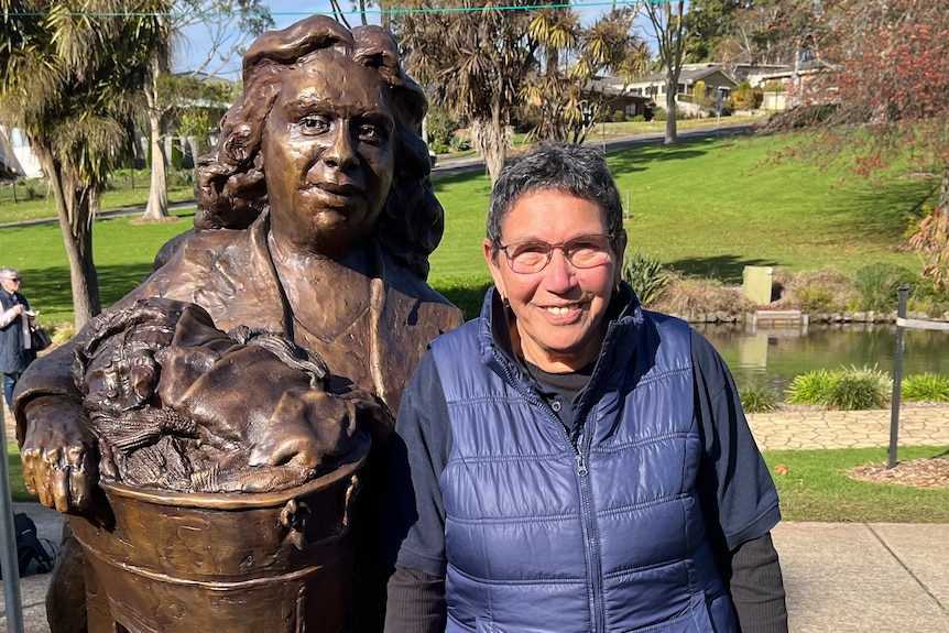 Aunty Cheryl has short hair, glasses and is wearing a blue puffer jacket next to statue of her mother holding a washing basket