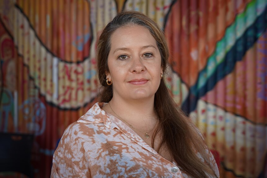 Woman in white and brown shirt looks at camera whilst standing in front of background with indigenous art