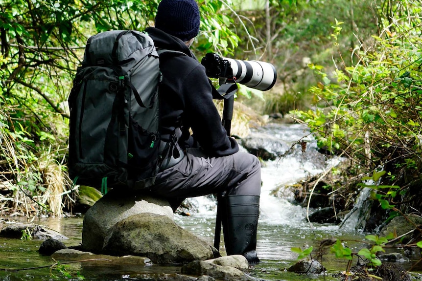 A man sitting on a river bank has his back to the camera while he holds a large camera.