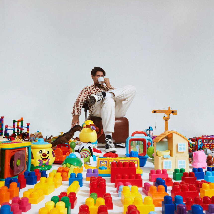 a man drinks from a cup as he sits on a stool inside a room littered with toys