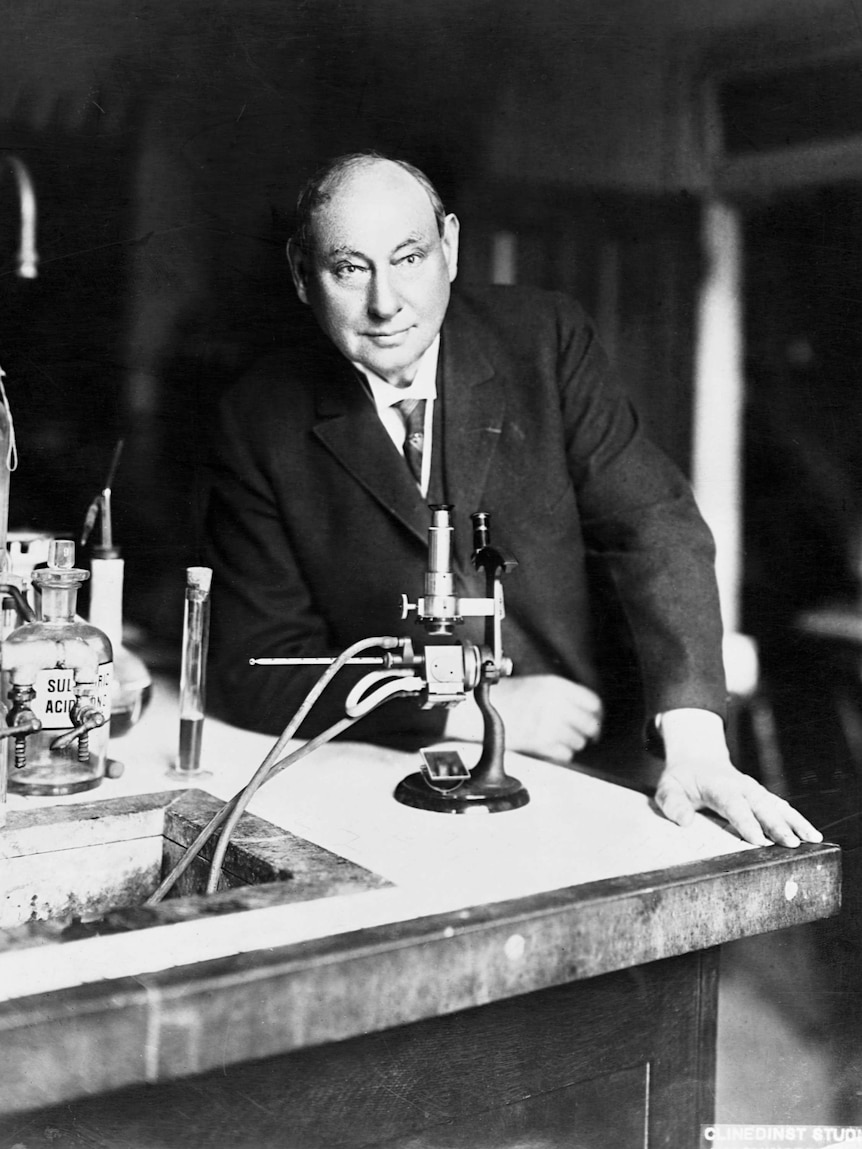 A black-and-white photo shows a bald man in a suit in a laboratory.