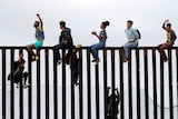 Central American migrants straddle the top of the border wall on the beach in San Diego. They are clapping and cheering.