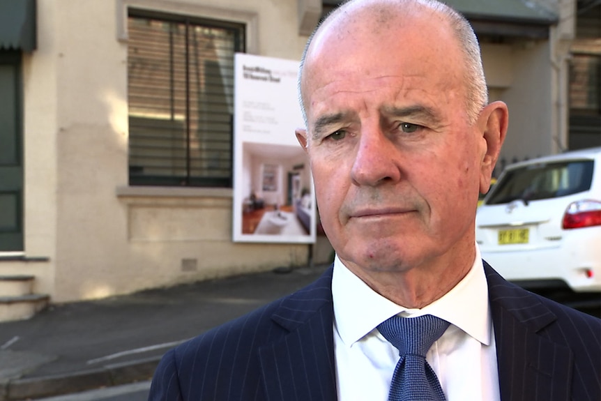 Malcolm Gunning stands outside a house with a For Sale sign on it