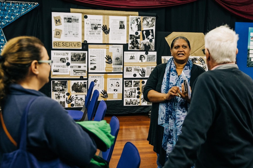 Indigenouse woman talking to grey haired man on right and women on left in front of display boards