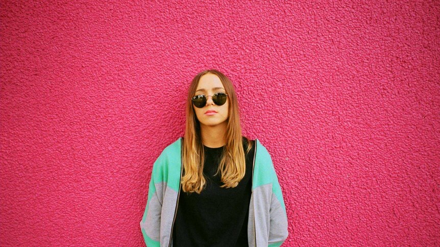 Nyxen standing in front of a pink wall, wearing sunnies and a sports jacket