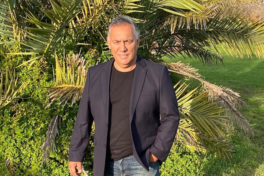 Wide shot of man wearing jeans and black shirt and suit jacket, standing on green lawn in front of a palm tree like plant