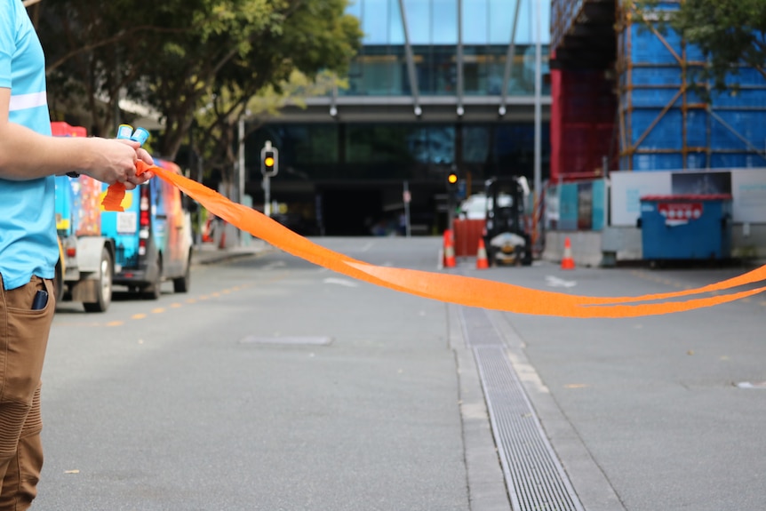 A person holds a party popper and a stream across an urban bitumen road, with traffic lights and construction in the background.