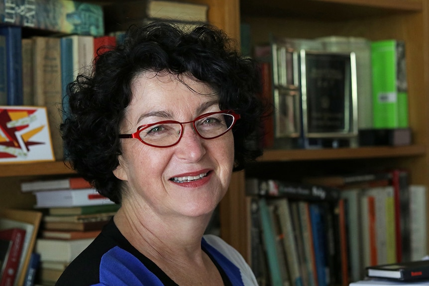 A woman wearing red glasses smiles at the camera, bookshelf in background.