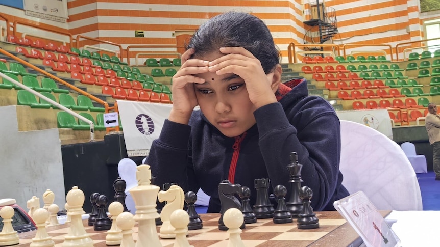 Young girl holds hands to forehead while looking at a chessboard. 