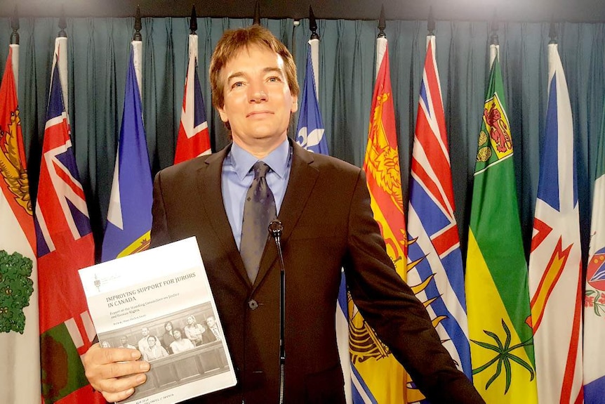 A man in a suit stands in front of a curtain and a row of flags, holding a document with the title Improving Support for Jurors