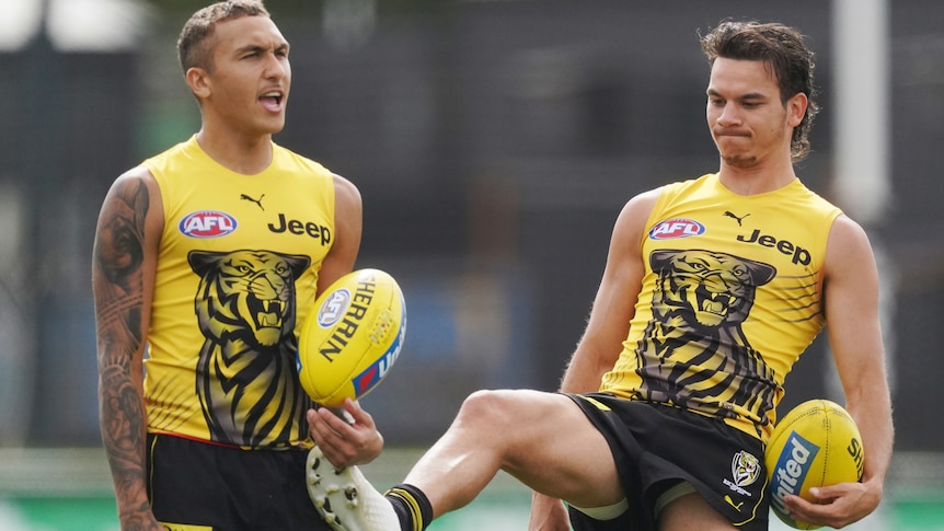 Shai Bolton holds a footy while Daniel Rioli tries to kick it out of his hand. They are wearing their training jerseys