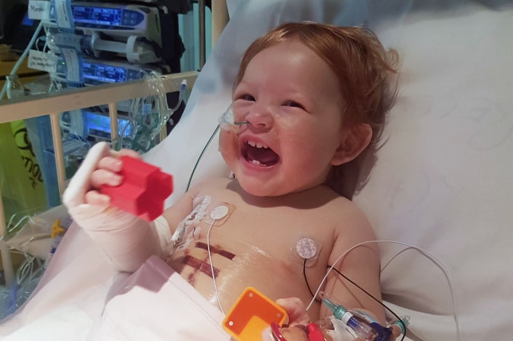 Mila, laughing, lies in a hospital bed with medical wires attached to her body.