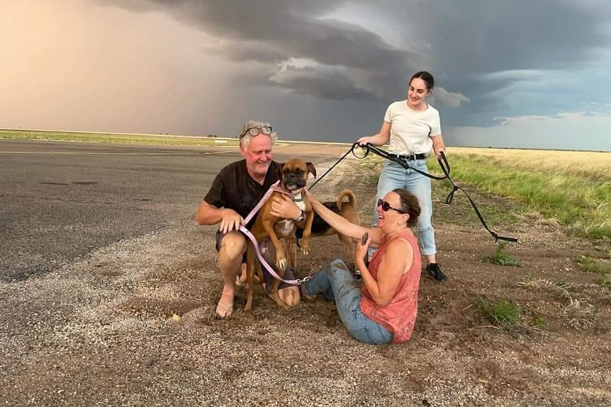 A man and two woman hold a brown and black Boxer dog underneath a dark sky next to an airport runway.