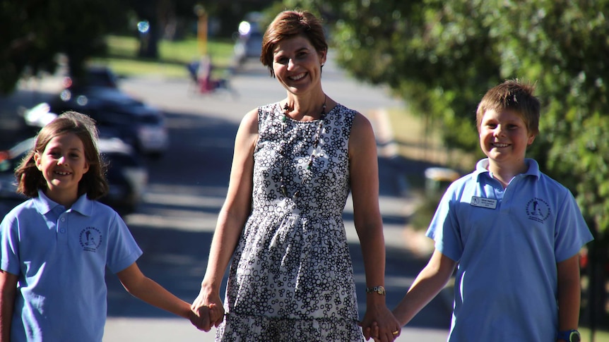 Kellie holds her children's hands and walks down the street. Green trees are visible in the background.