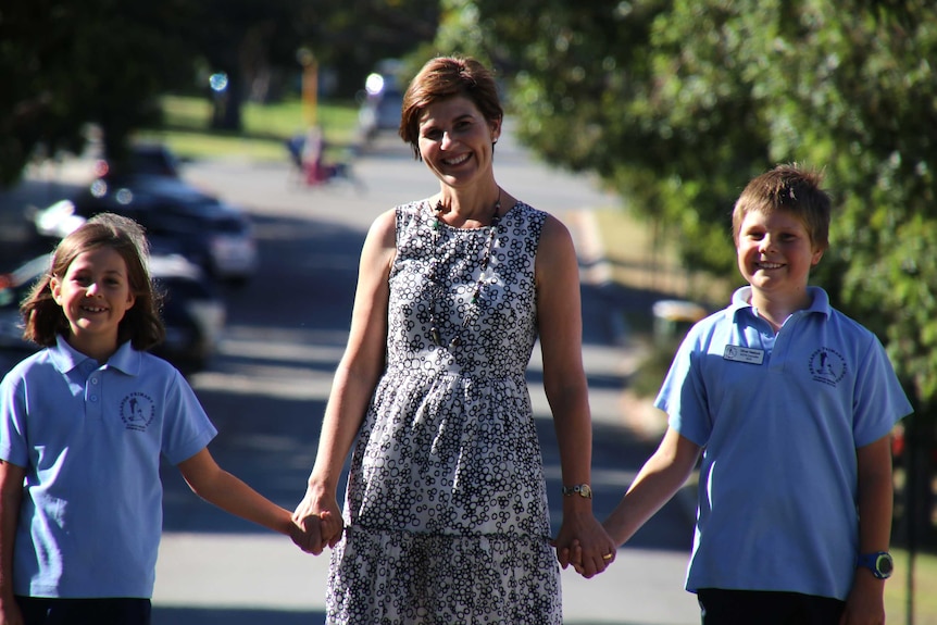 Kellie holds her children's hands and walks down the street. Green trees are visible in the background.