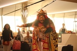 A person wearing cultural clothing including colorful robes, headwear and a mask performs a dance.