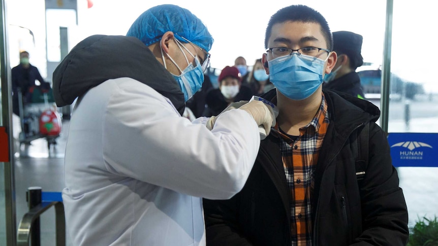 A man in white coat, gloves, masks and cap puts a thermometer to a young masked man's neck in an airport departure lounge.