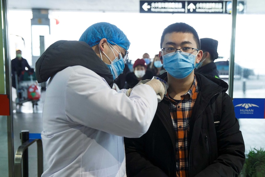 A man in white coat, gloves, masks and cap puts a thermometer to a young masked man's neck in an airport departure lounge.