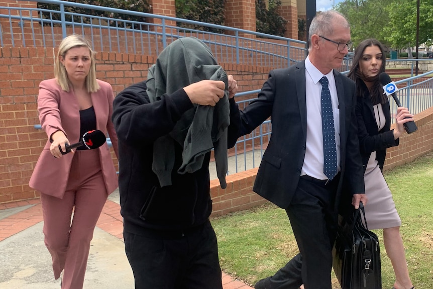 A man hides his face under a jumper as he leaves court surrounded by two reporters and his lawyer.