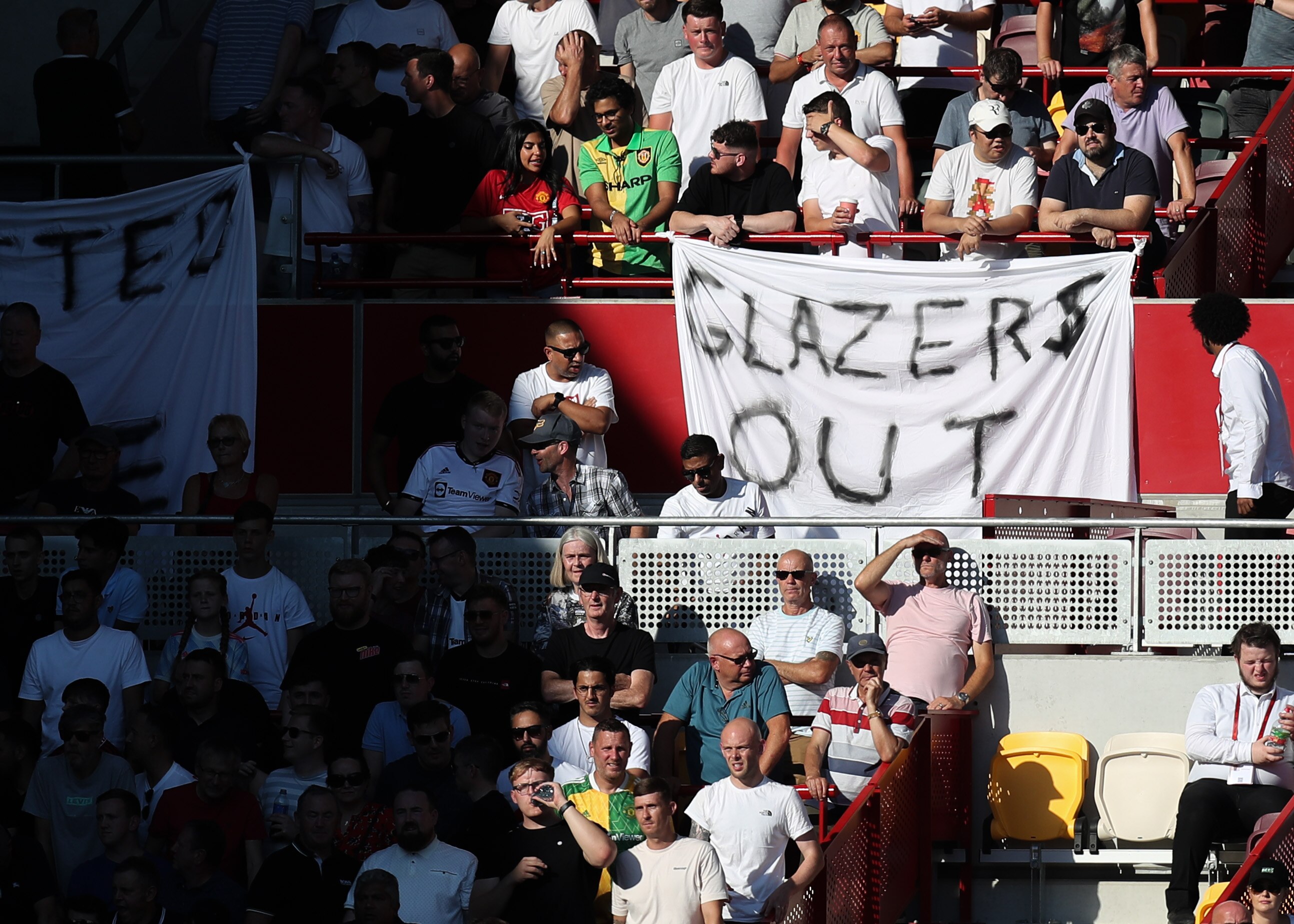 Football fans sit in a stand during a game with a banner saying 'Glazers Out'.
