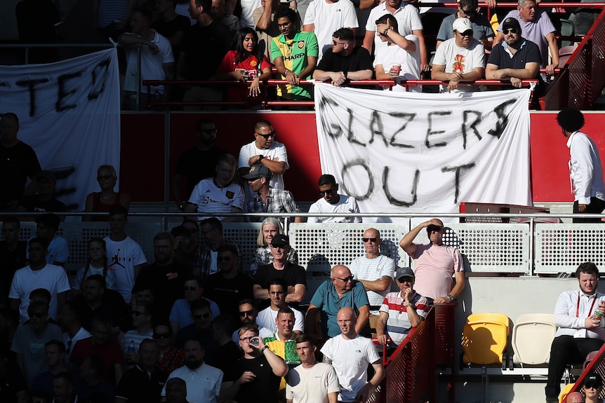 Football fans sit in a stand during a game with a banner saying 'Glazers Out'.