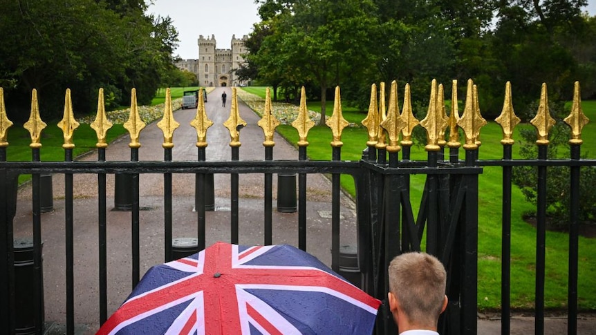 Members of the public, one with an umbrella with the Britain national flag on, stand outside Windsor Castle