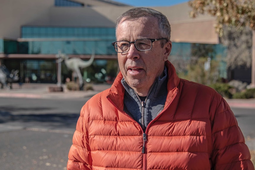 Spencer Lucas wear a puffy orange jacket, mid-speech, standing outside the museum