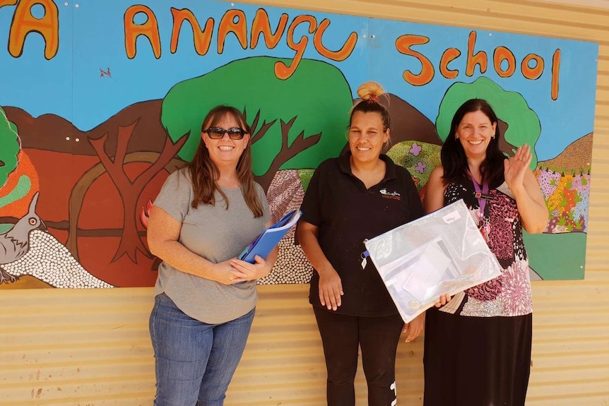 Three women standing in front of a painting with the words "Anangu School"